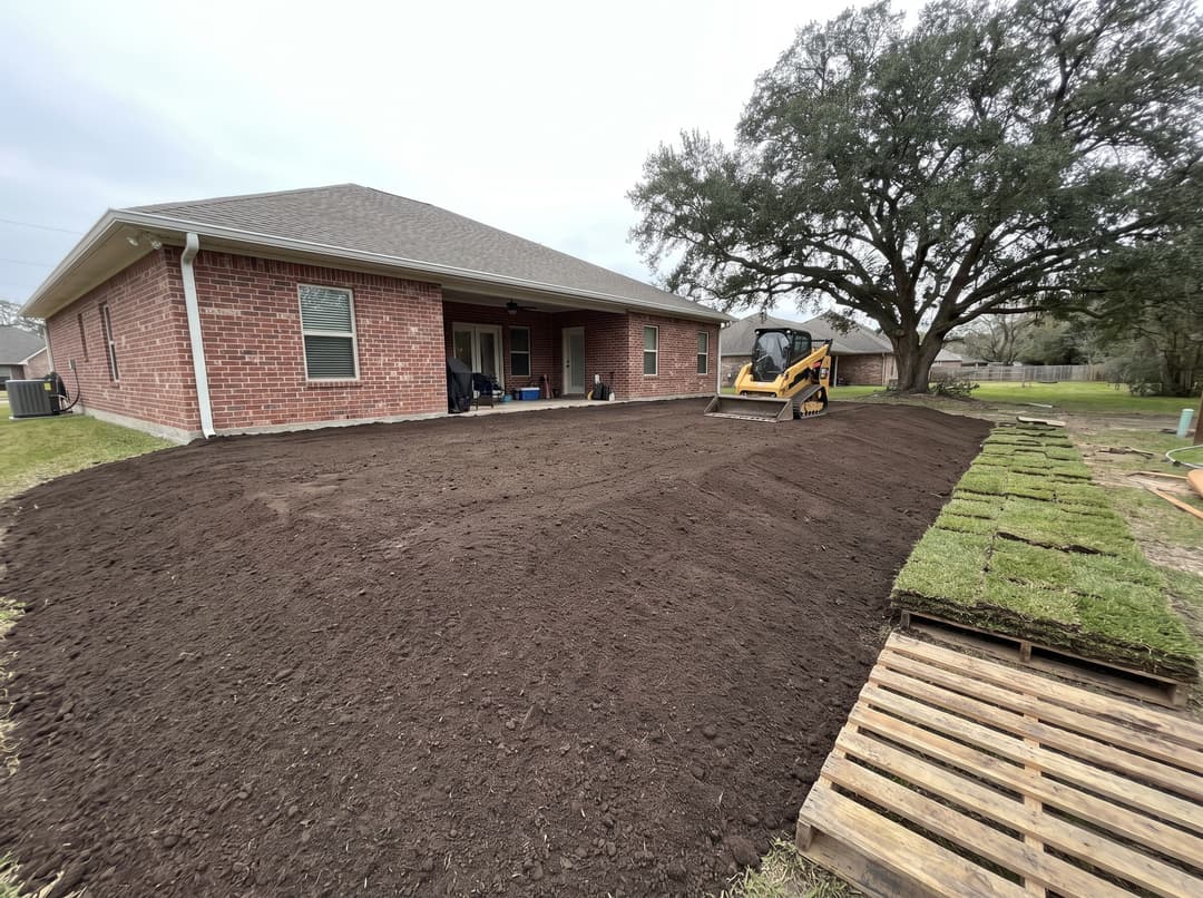 A backyard freshly regraded with smooth topsoil sloping away from a brick house, with a small loader and new sod nearby.